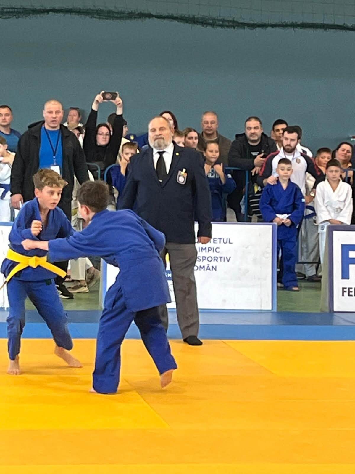 Doi copii în kimono albastru în timpul unui schimb pe tatami galben, arbitru în costum alături